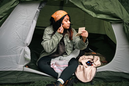 Femme assise dans une tente, appliquant du maquillage avec un miroir, portant un bandana et une veste verte.