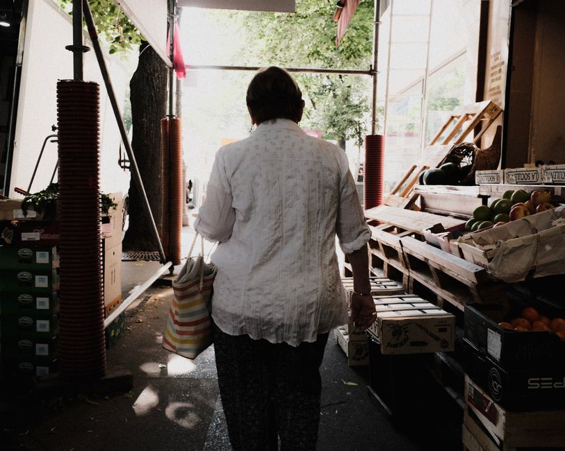 Femme portant une trousse de courses en tissu, marchant dans un marché de fruits et légumes.