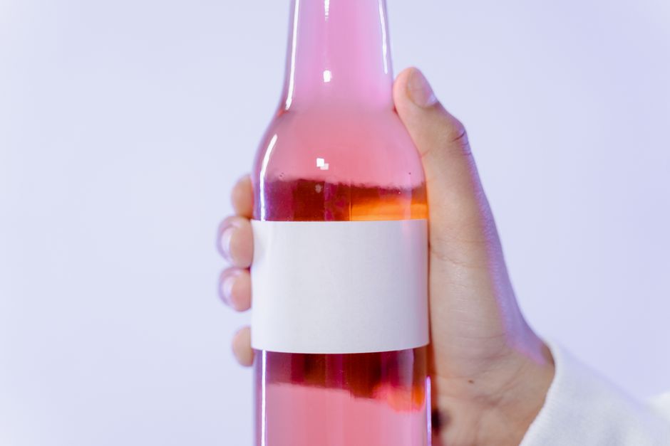 Main tenant une bouteille de boisson rose avec une étiquette blanche vide sur fond clair.