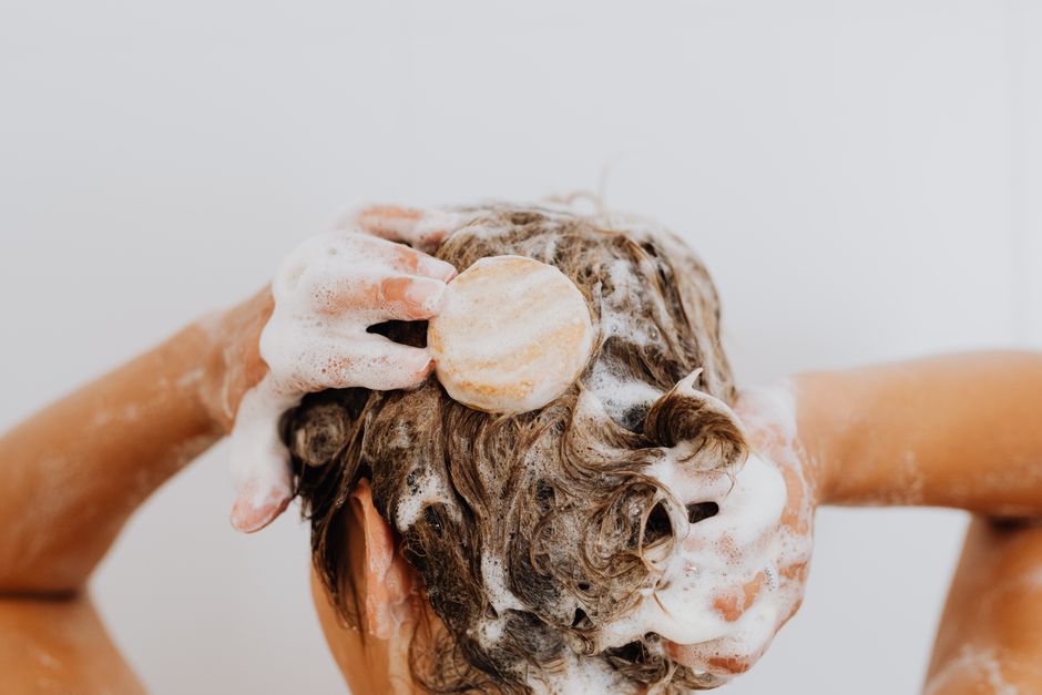 Femme appliquant un shampoing solide sur ses cheveux mouillés, mousse visible sur ses mains et cheveux.