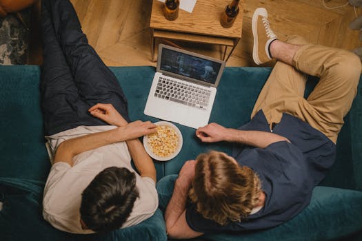 Deux personnes assises sur un canapé regardent un film sur un ordinateur portable, avec un bol de popcorn.