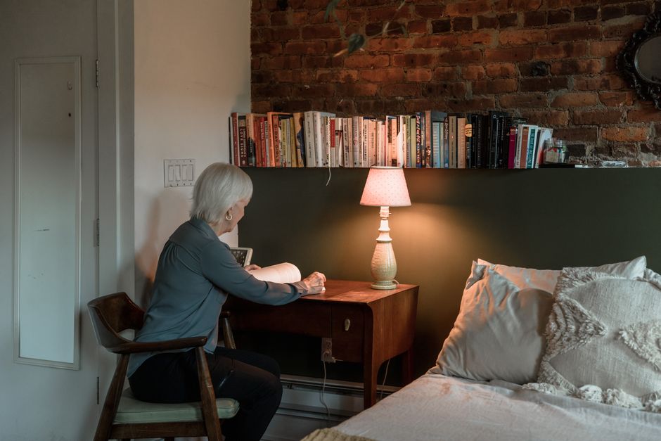 Femme âgée assise à une table de nuit, lisant un livre sous une lampe douce dans une chambre apaisante.