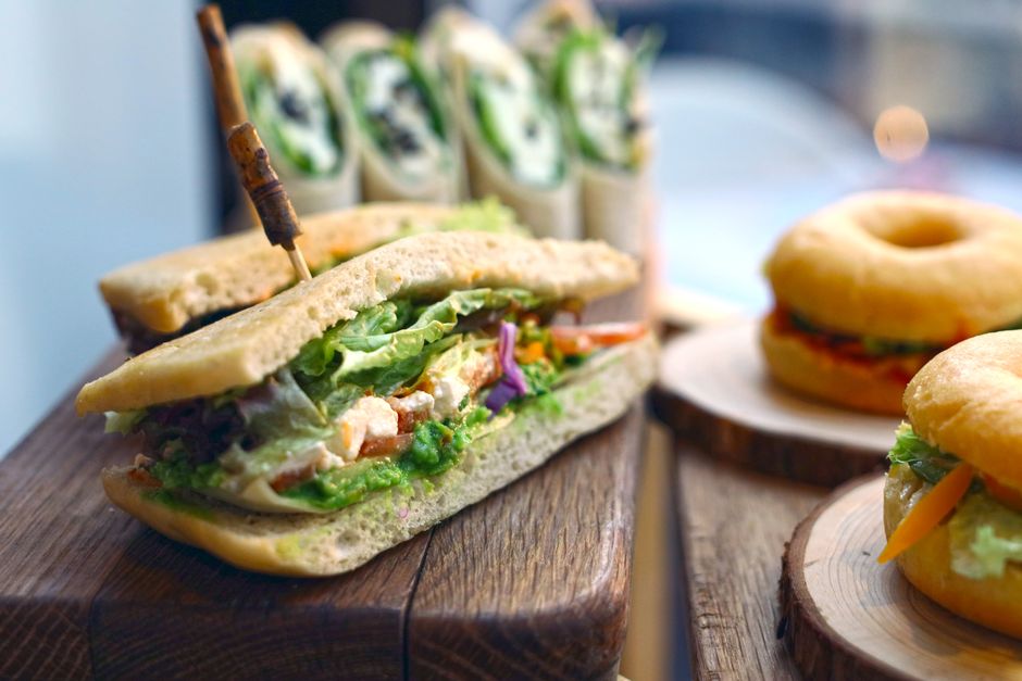 Sandwich garni de légumes et de poulet, avec des wraps et des donuts en arrière-plan sur une planche en bois.