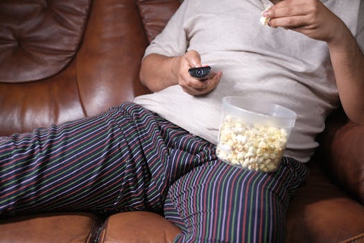 Homme assis sur un canapé en cuir, tenant une télécommande et un pot de pop-corn, vêtu de pyjama rayé.