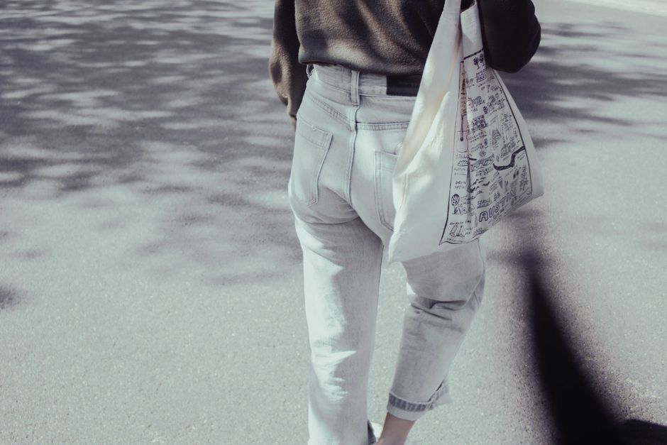 Femme marchant avec un sac en toile réutilisable, portant un pull et un jean, sur un chemin ensoleillé.