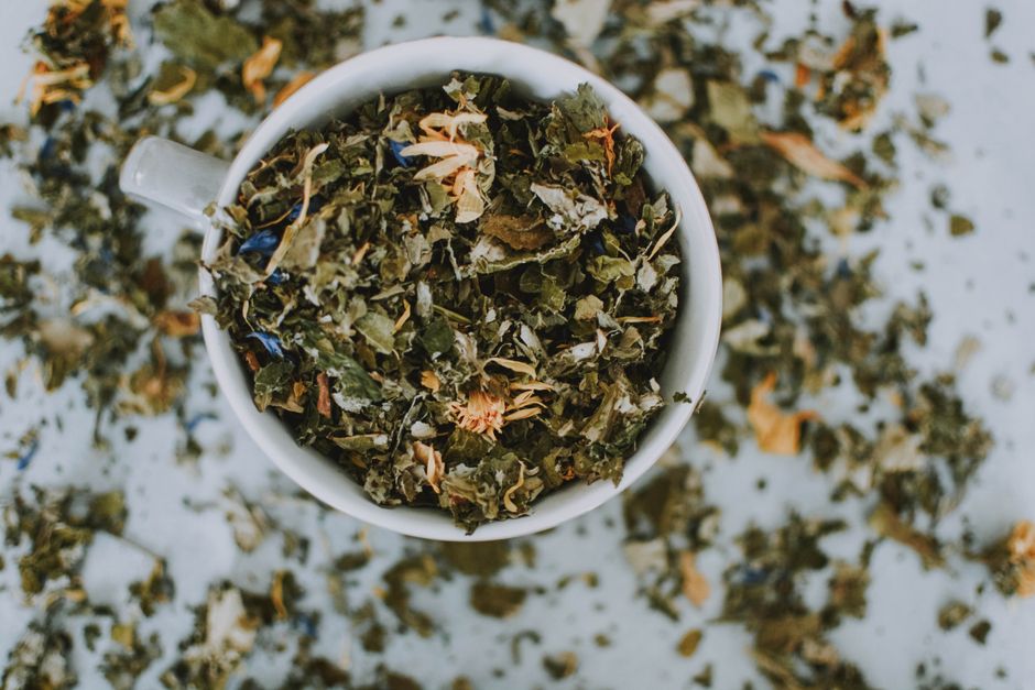 Tasse blanche remplie de mélange d'herbes et de fleurs séchées, entourée de feuilles éparpillées.