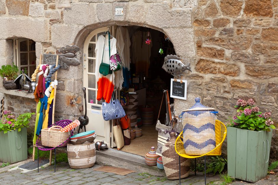 Boutique artisanale avec des paniers colorés et des vêtements suspendus à l'entrée, entourée de fleurs.